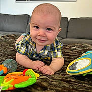 Archer is registered to the contest to win money with this photo: baby, child, smile, plaid_shirt, toy, blanket, couch, indoor, portrait, cute, playtime, infant, happy, face, person, furniture, home, closeup, lying_down, colorful