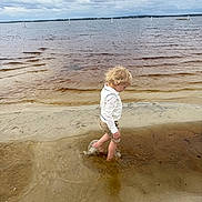 Hudson joined the competition — help win amazing prizes! child, toddler, beach, water, lake, sand, shoreline, waves, splash, barefoot, curly_hair, white_shirt, khaki_shorts, cloudy_sky, horizon, reflection, ripples, outdoor, walking, play
