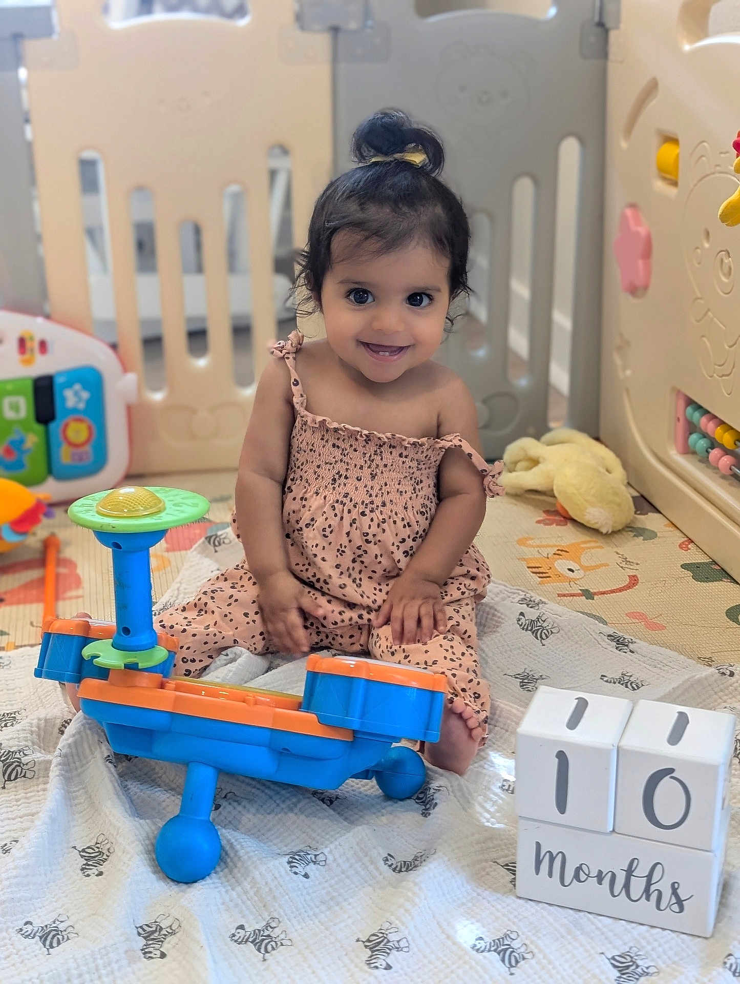 Shalev is registered to the contest to win money with this photo: baby, blanket, calendar, child, colorful, cute, dress, floor, furniture, happy, indoor, infant, months, plastic_toy, playpen, playtime, sitting, smile, topknot, toy
