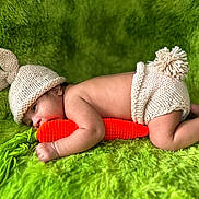 Julius is registered to the contest to win money with this photo: baby, infant, knitted_hat, knitted_diaper_cover, carrot, plush_carrot, green_blanket, fluffy_texture, cute, lying_down, baby_pose, soft_lighting, indoors, child, newborn, adorable, hat_with_ears, hand, face, skin