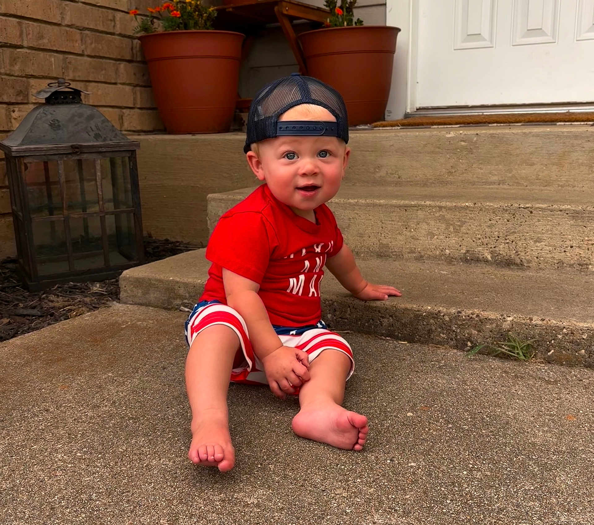 Kohen is registered to the contest to win money with this photo: toddler, child, baby, sitting, porch, concrete, cap, backwards_cap, shorts, red_shirt, barefoot, blue_eyes, potted_plants, lantern, outdoor, summer, curious, smiling, face, person