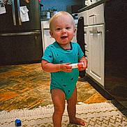 Kohen is registered to the contest to win money with this photo: toddler, child, baby, standing, kitchen, rug, floor, smiling, blue_onesie, barefoot, cabinet, refrigerator, toy, household, indoor, person, happy, home, blonde_hair, small_object