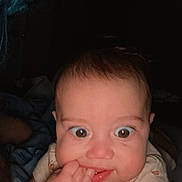 Flora is registered to the contest to win money with this photo: baby, infant, face, hand, fingers, curious, wide_eyes, chubby_cheeks, person, skin, hair, blue_hair, dark_background, clothing, cute, expression, portrait, indoor, close_up, child