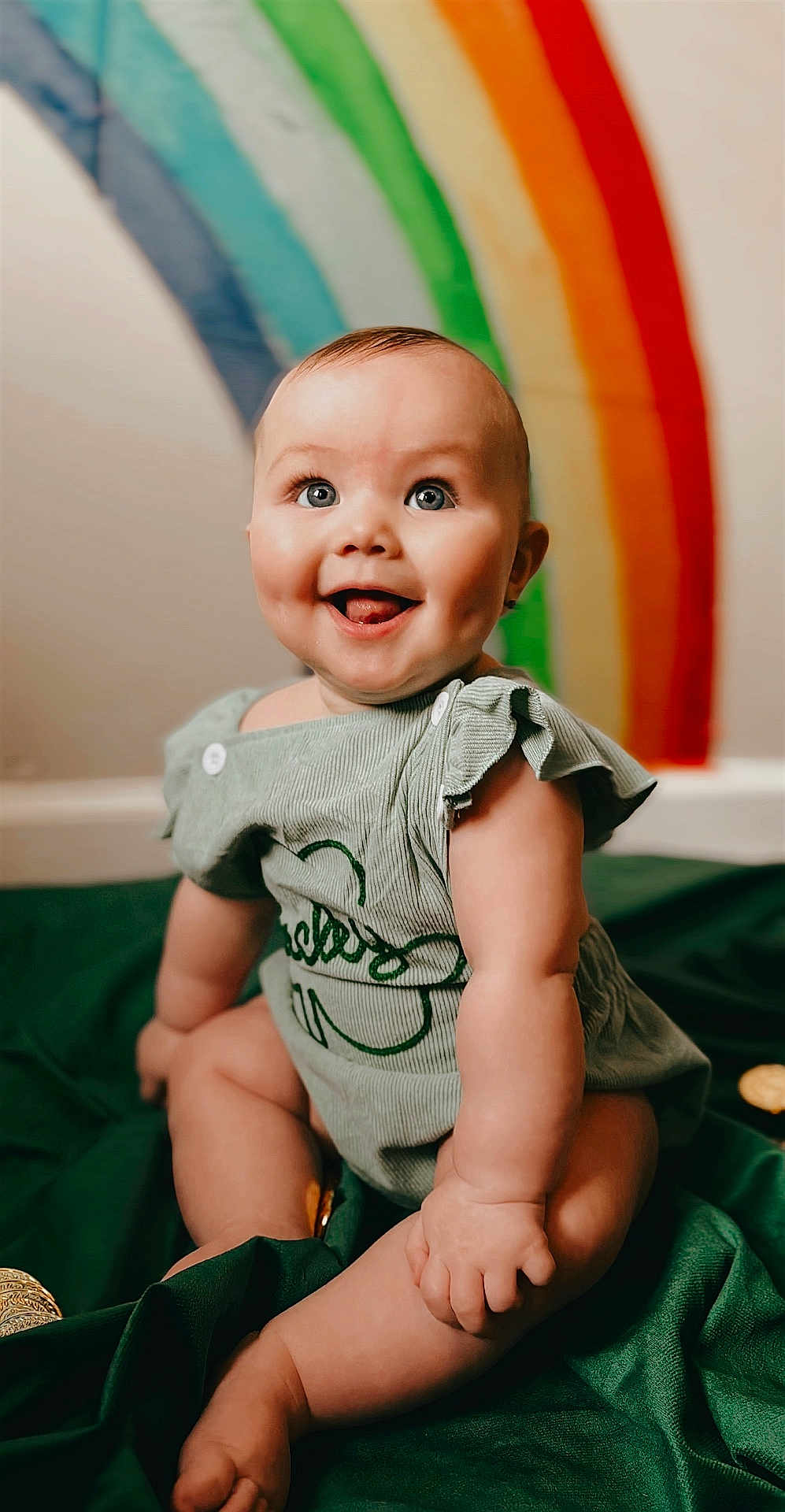 Alora joined the competition — help win amazing prizes! baby, infant, smiling, blue_eyes, dimples, green_outfit, rainbow, portrait, indoor, soft_lighting, sitting, blanket, cheeks, hands, legs, face, skin, button, happy, cute