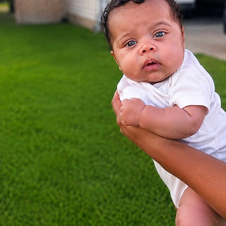 Carly is registered to the contest to win money with this photo: baby, infant, child, outdoor, grass, house, lawn, person, curious, holding, skin, white_clothing, hands, foot, portrait, daylight, suburban, expression, young, cute