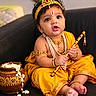 baby, barefoot, butter_pot, child, couch, flute, headpiece, indian_culture, indoor, infant, jewelry, musical_instrument, necklace, peacock_feather, portrait, sitting, smiling, traditional_clothing, wide_eyes, yellow_outfit