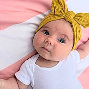 Jovie is registered to the contest to win money with this photo: baby, infant, headband, yellow, white_onesie, blanket, pink, white, face, cute, child, laying_down, portrait, soft_texture, newborn, expression, skin, clothing, indoors, closeup