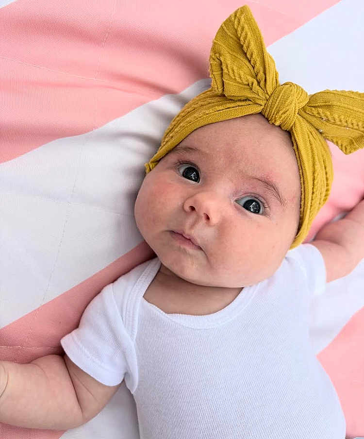 Jovie is registered to the contest to win money with this photo: baby, infant, headband, yellow, white_onesie, blanket, pink, white, face, cute, child, laying_down, portrait, soft_texture, newborn, expression, skin, clothing, indoors, closeup