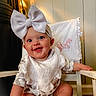baby, bow_headband, smile, chair, bib, infant, happy, indoor, person, pink_text, pillow, patterned_clothing, headwear, cute, child, skin, foot, hand, wood_floor, white_furniture