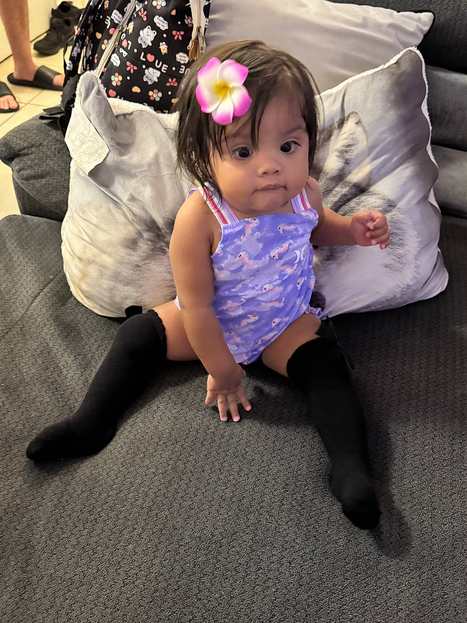 backpack, black_socks, carpet, child, couch, cute, face, flower, furniture, hair_accessory, home, indoor, legs, person, pillow, purple_outfit, sitting, smiling, toddler, young_child