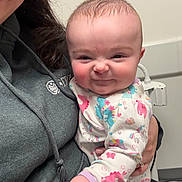 Octavia is registered to the contest to win money with this photo: baby, infant, child, smiling, scrunched_face, onesie, unicorn_print, parent, holding, indoor, clinic, portrait, close_up, hand, eyeglasses, hoodie, rosy_cheeks, playful_expression, cute, warm_clothing