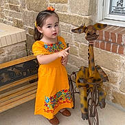 Camellia De Luna is registered to the contest to win money with this photo: child, girl, orange_dress, floral_pattern, pointing, metal_sculpture, giraffe, tricycle, bench, stone_wall, brick, outdoor, curious, playful, innocence, footwear, hair_accessory, decor, daylight, person