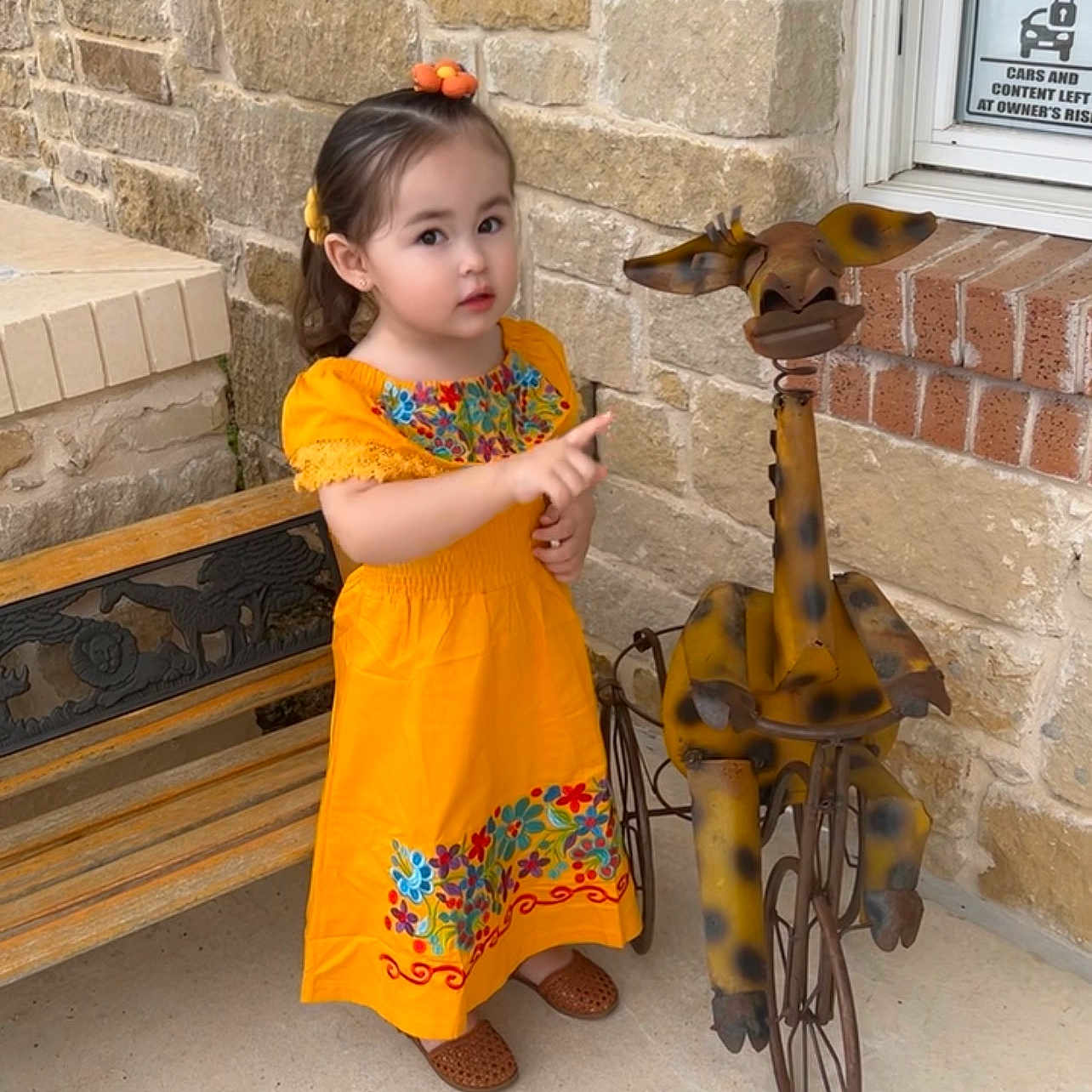 Camellia De Luna is registered to the contest to win money with this photo: child, girl, orange_dress, floral_pattern, pointing, metal_sculpture, giraffe, tricycle, bench, stone_wall, brick, outdoor, curious, playful, innocence, footwear, hair_accessory, decor, daylight, person