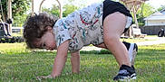 Oliver is registered to the contest to win money with this photo: toddler, child, grass, outdoor, playground, shoes, curly_hair, dinosaur_shirt, shorts, sunny_day, park, exploring, crawling, play_equipment, bench, tree, person, nature, summer, fun