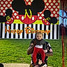 Carson joined the competition — help win amazing prizes! adult, baby, banner, birthday, celebration, child, dark_jacket, daylight, floral_shirt, green_grass, holding, mickey_mouse, outdoor, party, polka_dots, red_pants, smile, streamers, stripes, trees