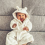 Phoebe is registered to the contest to win money with this photo: baby, child, smiling, hooded_robe, white_clothing, sitting, couch, furniture, happy, cute, indoors, portrait, cozy, plush, infant, face, expression, soft, warm, comfortable