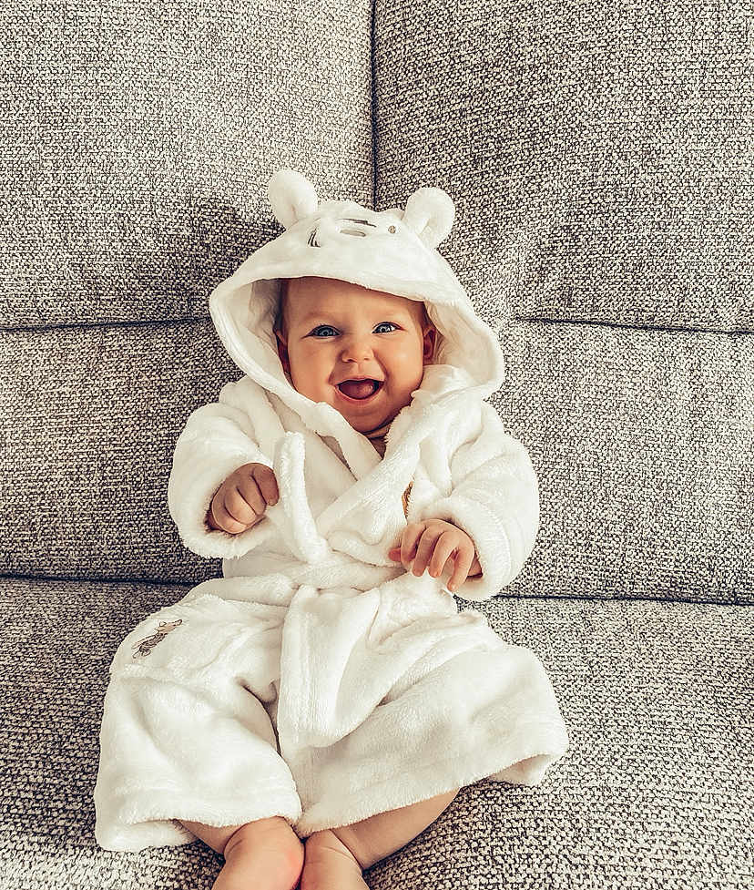 Phoebe is registered to the contest to win money with this photo: baby, child, smiling, hooded_robe, white_clothing, sitting, couch, furniture, happy, cute, indoors, portrait, cozy, plush, infant, face, expression, soft, warm, comfortable