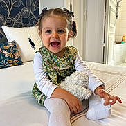 Salomé a rejoint le concours — aidez-le/la à gagner de superbes lots ! toddler, child, smiling, pigtails, bows, green_dress, white_shirt, white_tights, stuffed_animal, bed, pillow, floral_pattern, indoor, happy, cute, portrait, person, home, cozy, playful