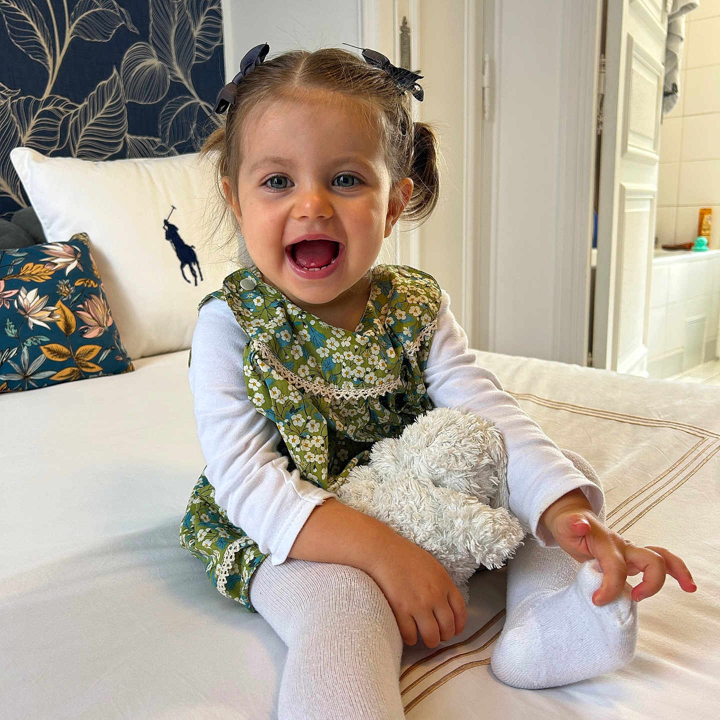 Salomé a rejoint le concours — aidez-le/la à gagner de superbes lots ! bed, bows, child, cozy, cute, floral_pattern, green_dress, happy, home, indoor, person, pigtails, pillow, playful, portrait, smiling, stuffed_animal, toddler, white_shirt, white_tights