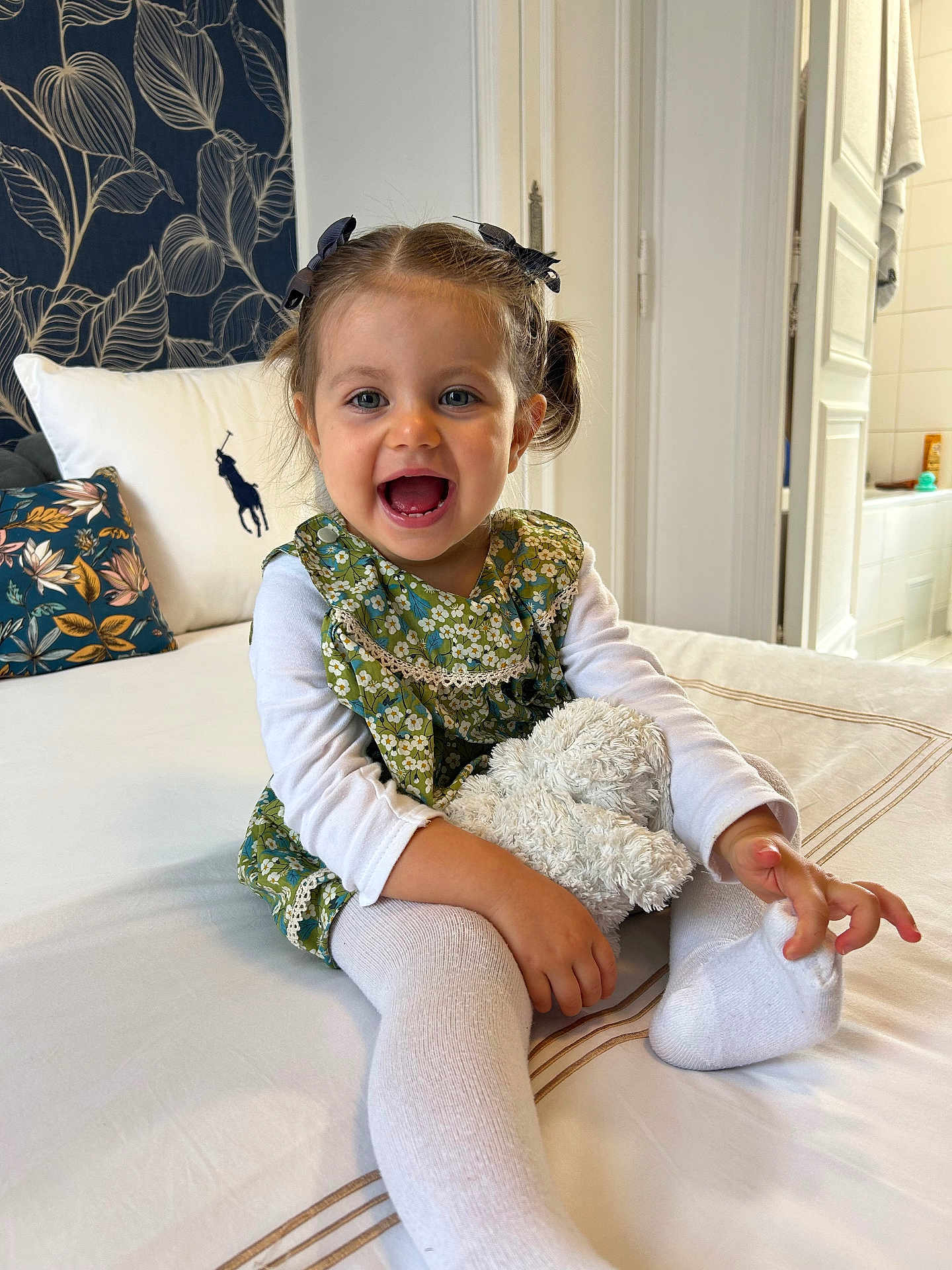 Salomé a rejoint le concours — aidez-le/la à gagner de superbes lots ! toddler, child, smiling, pigtails, bows, green_dress, white_shirt, white_tights, stuffed_animal, bed, pillow, floral_pattern, indoor, happy, cute, portrait, person, home, cozy, playful
