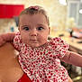 baby, child, infant, face, portrait, pink_bow, floral_dress, person, curious, eyes, cheeks, arm, red_clothing, blurred_background, indoor, holding, skin, head, cute, closeup