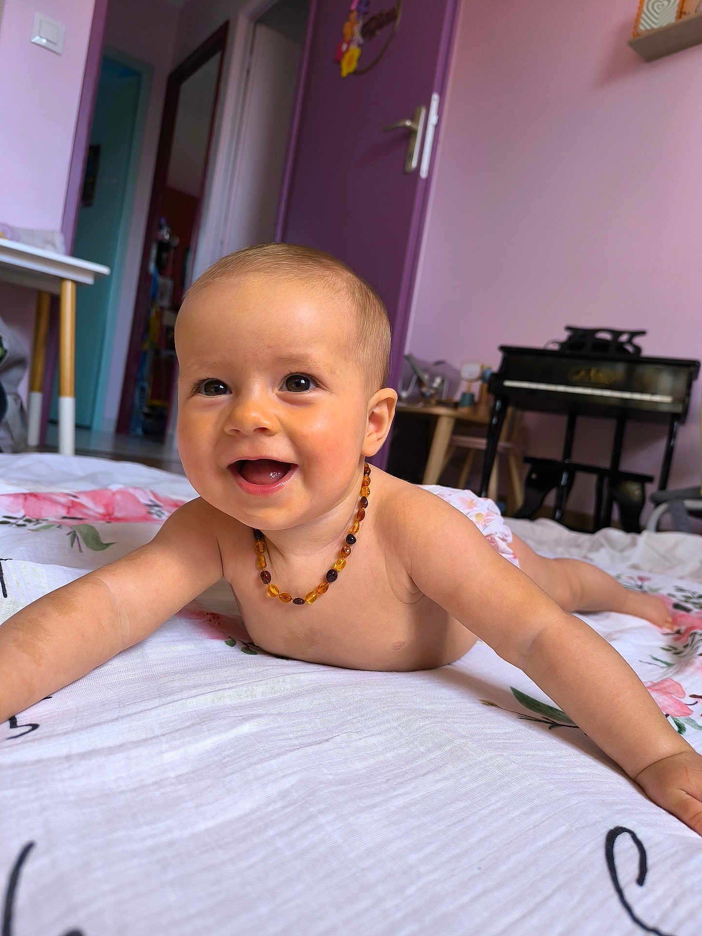 Yüna a rejoint le concours — aidez-le/la à gagner de superbes lots ! baby, infant, smiling, lying_down, amber_necklace, blanket, floral_pattern, indoor, room, purple_wall, door, toy_piano, furniture, child, happy, cute, portrait, skin, person, floor