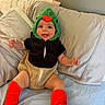 Maverick joined the competition — help win amazing prizes! baby, child, dinosaur_costume, costume, green, red, orange, smiling, sitting, bed, pillows, bedroom, indoor, portrait, face, infant, clothing, leg_warmers, happy, photography