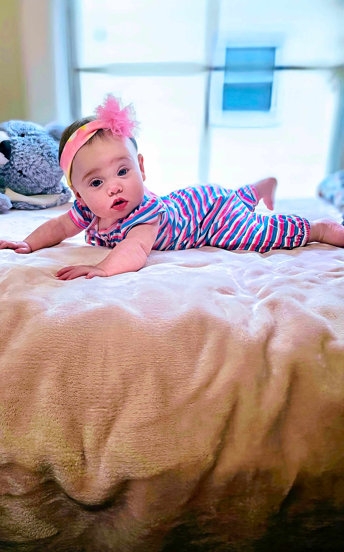 Ara is registered to the contest to win money with this photo: baby, infant, bed, blanket, striped_clothing, headband, pink, plush_toy, window, indoor, child, cute, portrait, soft_texture, lying_down, face, expression, curious, home, natural_light