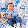 baby, infant, child, smiling, sitting, portrait, blue_theme, teddy_bear, stuffed_animal, balloon, crib, blanket, plush_toy, baby_clothing, jeans, sneakers, nursery, birthday_sign, indoor, happy