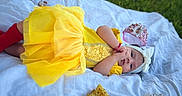 Meredith is registered to the contest to win money with this photo: baby, yellow_dress, headband, blanket, outdoor, grass, toy, tiara, princess, child, cute, lying_down, infant, playtime, soft_fabric, portrait, smiling, red_bracelet, decor, person