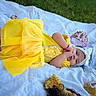 baby, yellow_dress, headband, blanket, outdoor, grass, toy, tiara, princess, child, cute, lying_down, infant, playtime, soft_fabric, portrait, smiling, red_bracelet, decor, person