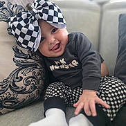 Rayven joined the competition — help win amazing prizes! baby, child, smiling, tongue_out, bow, checkered_pattern, pants, long_sleeve_shirt, couch, pillow, socks, indoor, cute, happy, portrait, seated, clothing, face, person, playful