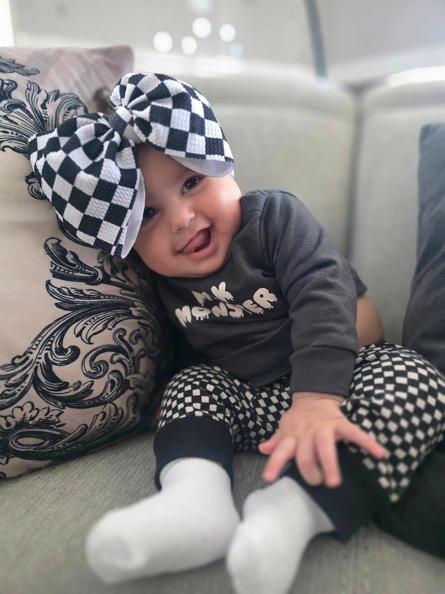 Rayven joined the competition — help win amazing prizes! baby, child, smiling, tongue_out, bow, checkered_pattern, pants, long_sleeve_shirt, couch, pillow, socks, indoor, cute, happy, portrait, seated, clothing, face, person, playful