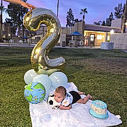 Christopher joined the competition — help win amazing prizes! baby, blanket, balloon, number_2, grass, cake, soccer_ball, hand, outdoor, evening, tree, palm_tree, celebration, party, blue_sky, building, fence, person, lawn, toddler