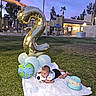 baby, blanket, balloon, number_2, grass, cake, soccer_ball, hand, outdoor, evening, tree, palm_tree, celebration, party, blue_sky, building, fence, person, lawn, toddler