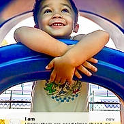 Elias is registered to the contest to win money with this photo: child, smiling, playground, outdoor, blue_bar, crossed_arms, portrait, happy, face, teeth, curly_hair, toddler, clothing, t_shirt, play_equipment, colorful, sunlight, fence, social_media, text_overlay