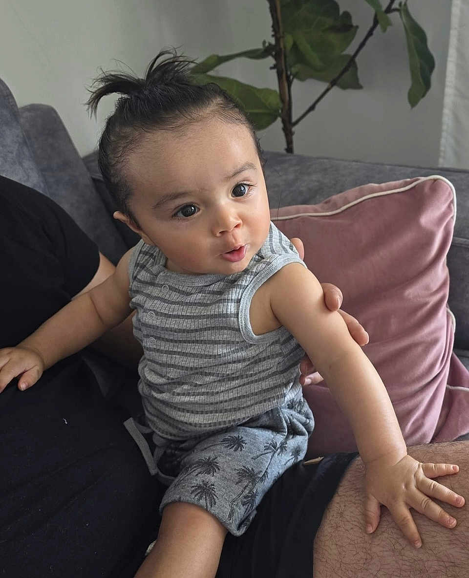 Chalom a rejoint le concours — aidez-le/la à gagner de superbes lots ! casual_clothing, child, couch, curious, expression, furniture, hair_tied, hand, home, indoor, lap, palm_tree_pattern, person, pillow, plant, relaxed, shorts, sitting, striped_shirt, toddler
