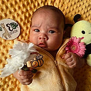 Kai is registered to the contest to win money with this photo: baby, infant, flower, plush_toy, bee, yellow_blanket, milestone_card, cute, holding, soft_texture, baby_clothes, portrait, child, indoors, expressive_face, newborn, hand, pink_flower, white_flower, lying_down