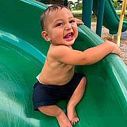 Ayce is registered to the contest to win money with this photo: toddler, child, smiling, playground, slide, green_slide, outdoor, park, happy, bare_chest, shorts, feet, play, fun, young_child, daylight, nature, grass, trees, summer