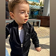 Milann a rejoint le concours — aidez-le/la à gagner de superbes lots ! toddler, child, outdoor, sunlight, jacket, black_clothing, white_shirt, curious, person, urban, bench, pavement, building, daylight, portrait, casual_wear, young_child, face, hair, expression