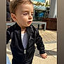 toddler, child, outdoor, sunlight, jacket, black_clothing, white_shirt, curious, person, urban, bench, pavement, building, daylight, portrait, casual_wear, young_child, face, hair, expression