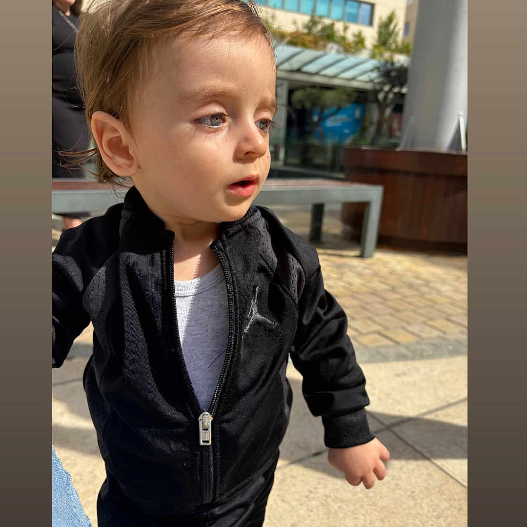 Milann a rejoint le concours — aidez-le/la à gagner de superbes lots ! bench, black_clothing, building, casual_wear, child, curious, daylight, expression, face, hair, jacket, outdoor, pavement, person, portrait, sunlight, toddler, urban, white_shirt, young_child