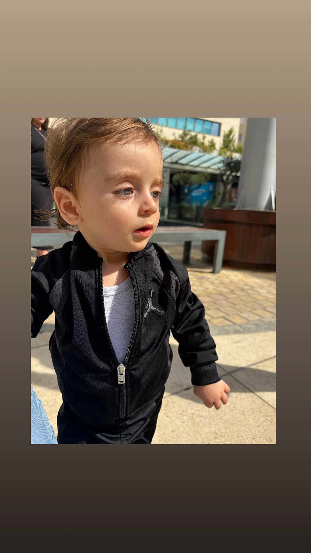 Milann a rejoint le concours — aidez-le/la à gagner de superbes lots ! toddler, child, outdoor, sunlight, jacket, black_clothing, white_shirt, curious, person, urban, bench, pavement, building, daylight, portrait, casual_wear, young_child, face, hair, expression
