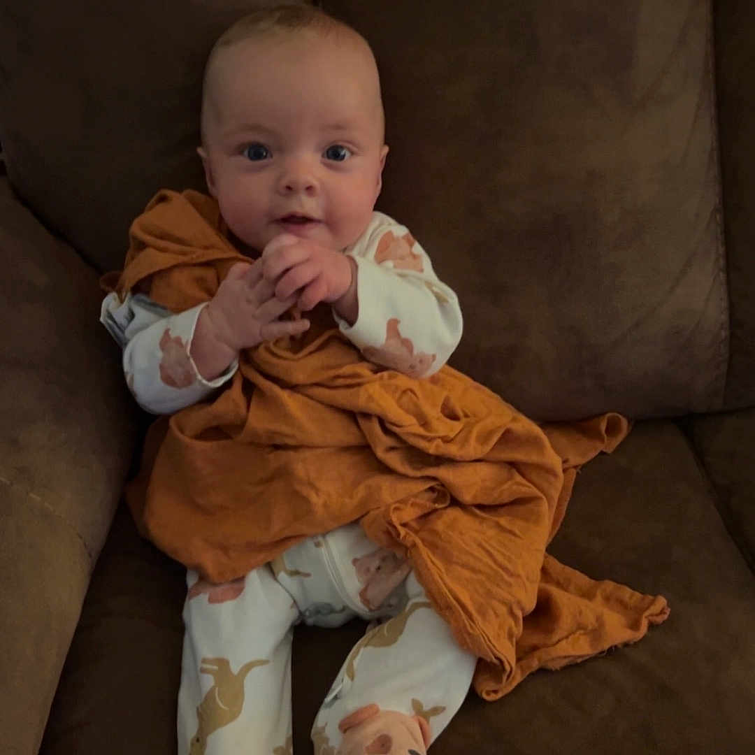 Merliah joined the competition — help win amazing prizes! baby, infant, child, couch, blanket, pajamas, animal_print, footwear, face, hands, sitting, indoors, cute, cozy, expression, clothing, furniture, brown, orange, portrait