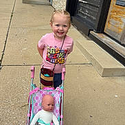 Eliana is registered to the contest to win money with this photo: child, girl, smiling, toy_stroller, doll, sidewalk, braided_hair, pink, shirt, casual_clothing, outdoor, daylight, storefront, urban, person, play, happy, small_child, street, accessory
