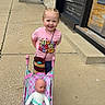 accessory, braided_hair, casual_clothing, child, daylight, doll, girl, happy, outdoor, person, pink, play, shirt, sidewalk, small_child, smiling, storefront, street, toy_stroller, urban