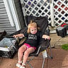 toddler, child, chair, outdoor, pavement, lattice_fence, black_shirt, pink_shorts, footwear, sandals, hair_tie, plastic_crate, toolbox, sunlight, relaxing, summer, backyard, person, small_pony_tail, concrete
