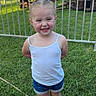 child, smiling, grass, amusement_ride, outdoor, summer, tank_top, denim_shorts, white_sneakers, fence, playground, happy, young_girl, casual_clothing, daylight, greenery, fun, recreation, portrait, park