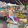 amusement_ride, background, blue_jeans, carnival, child, colorful, excitement, fair, fun, lights, metal_railings, motion_blur, motorcycle_ride, outdoor, playful, purple_canopy, smiling, white_shoes, yellow_shirt, young_child