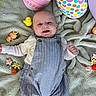 baby, smiling, blanket, overalls, easter_eggs, toys, flowers, indoor, child, cute, happy, playful, colorful, patterned, clothing, portrait, infant, soft_texture, decorations, lying_down